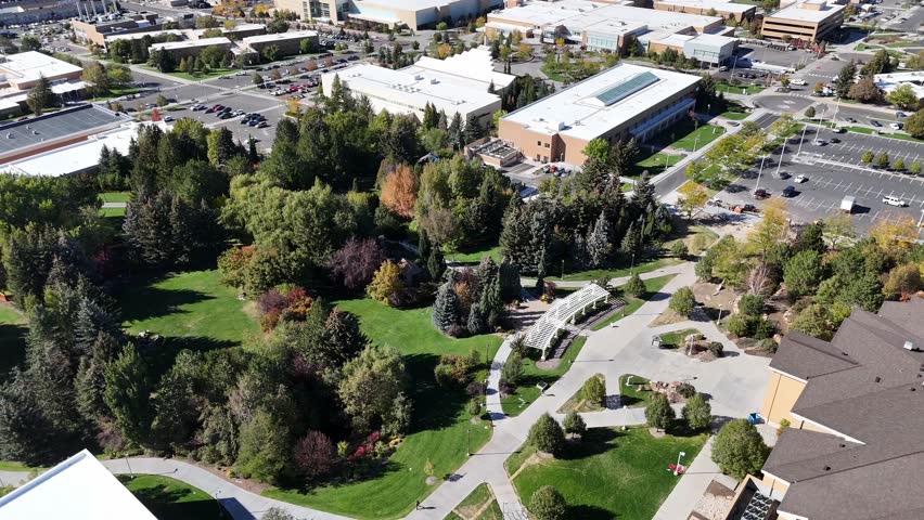 An Aerial view of a city park with green lawns, tall trees, parking lot and nearby commercial buildings on a sunny day in Boise, Idaho