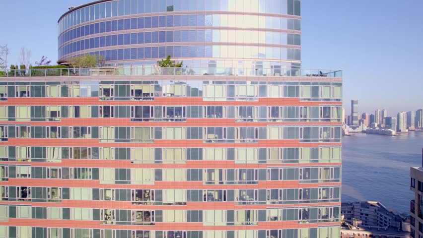 A close-up drone view of a luxury residential tower in Battery Park, New York, with a curved glass facade facing the Hudson River and skyline