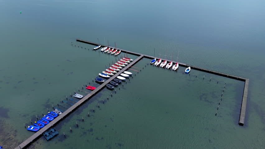 A magnificent high-angle aerial photograph capturing a drone overview of a calm harbor station for recreational boating and water sports. The composition centers on a geometric, T-shaped wooden jetty and dock system floating on the clear, green-tinted water of a tranquil lake, lagoon, or sea bay. Neatly moored along the wooden planks are rows of various watercraft, including several small white sailing yachts with masts, bright red and blue dinghy boats, small motorboats, and plastic pedal boats. The glassy surface of the water reveals subtle underwater sand patches and seaweed near the shallow shore. The soft, even daylight enhances the crisp colors of the vessels, perfectly highlighting the peaceful atmosphere, coastal leisure lifestyle, sailing school infrastructure, and outdoor summer recreation typical of European holiday lakes and maritime sports clubs.