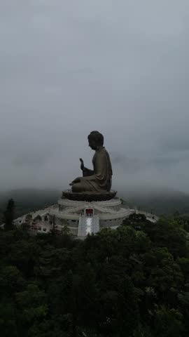Majestic Tian Tan Big Buddha statue sitting on a lotus throne on Lantau Island in Hong Kong, filmed from above with a drone flying over the monument on a very foggy and moody day.