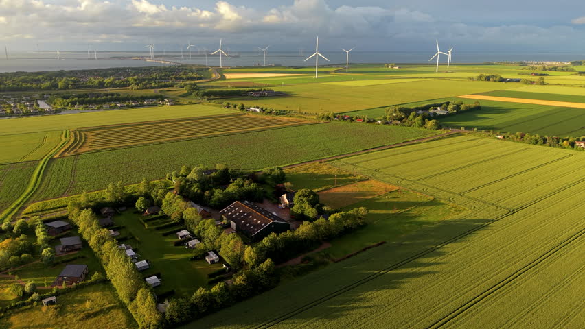 A magnificent high-angle aerial photograph capturing a drone overview of a scenic European countryside during the late afternoon golden hour. The composition tightly focuses on a traditional farmhouse estate featuring a large dark barn roof, private gardens, and an adjacent small camping ground filled with white caravans and small holiday chalets. The entire property is enveloped by mature green trees and nestled within an expansive patchwork of vibrant green agricultural fields showing distinct tractor track lines. In the background, a massive onshore wind farm with numerous tall white wind turbines lines the sea edge, with a prominent coastal bridge or storm surge barrier visible on the horizon. A dramatic sky with sunlit cumulus clouds casts long, soft shadows across the flat plains. This stunning panoramic landscape beautifully documents rural hospitality, green energy integration, and peaceful Dutch environmental scenery.