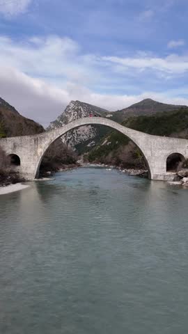 A cinematic, vertical drone footage beneath the historic Plaka Arch Bridge, spans the Arachthos River in the Epirus region of Greece