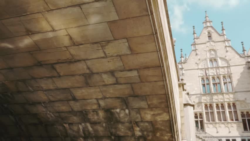 A low angle view taken from a passing boat under stone bridge across canal, showcasing the ornate architectures under clear skies in Ghent, Belgium