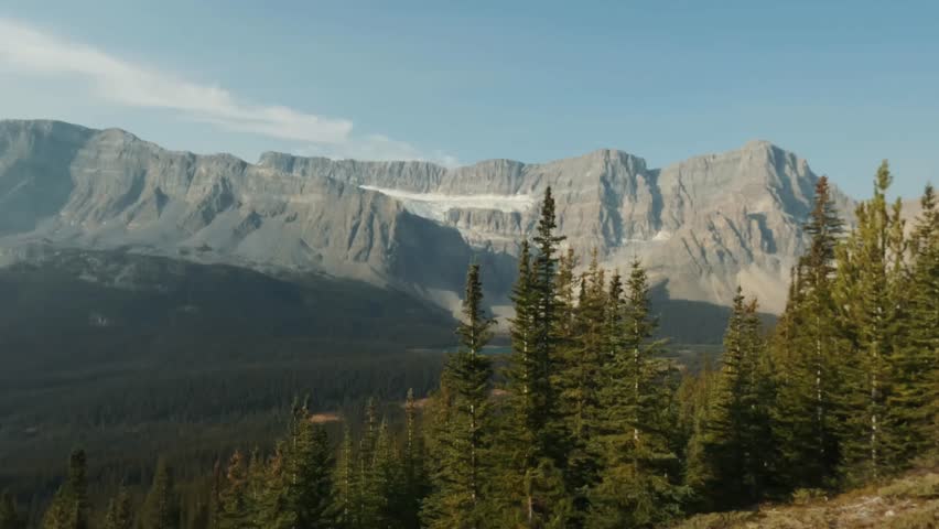 A view of the Rocky Mountains in Banff National Park, Alberta, dotted with patches of snow and framed by lush evergreen forests under a clear blue sky