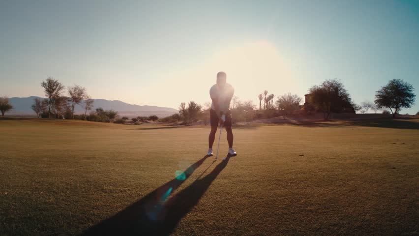 Golfer hits wedge pitch at golden hour in the desert, silhouetted against a sun flare through filmed in slow motion,