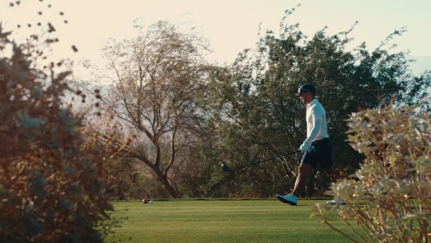 A lone golfer approaches his tee shot, framed between desert bushes at backlit golden hour, filmed in slow motion