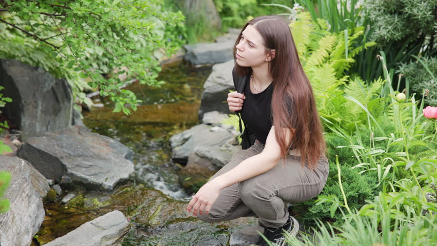 Young woman crouching beside garden stream inspecting water and plants in lush backyard