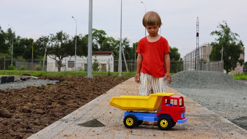 A four-year-old child in beige boots and shorts plays with a toy dump truck with a yellow semi-elliptical bed. The boy is playing construction with toy construction equipment, unloading the truck