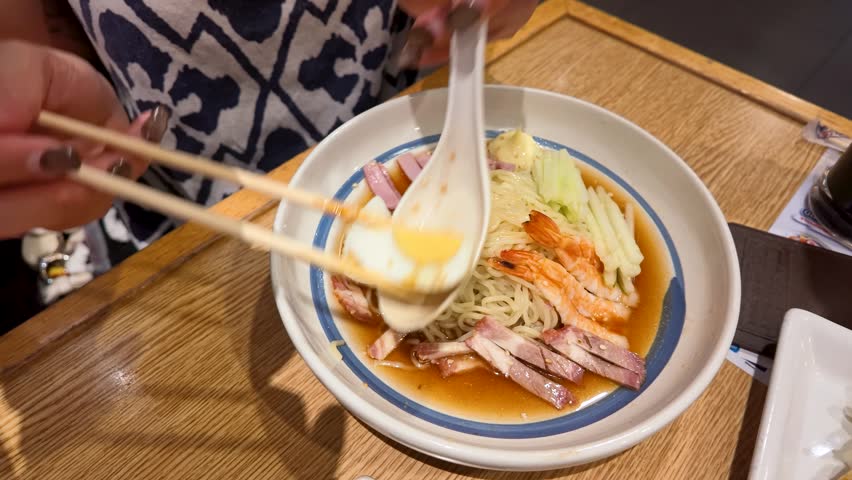 Overhead view of a diner mixing cold ramen noodles with shrimp and egg using chopsticks.