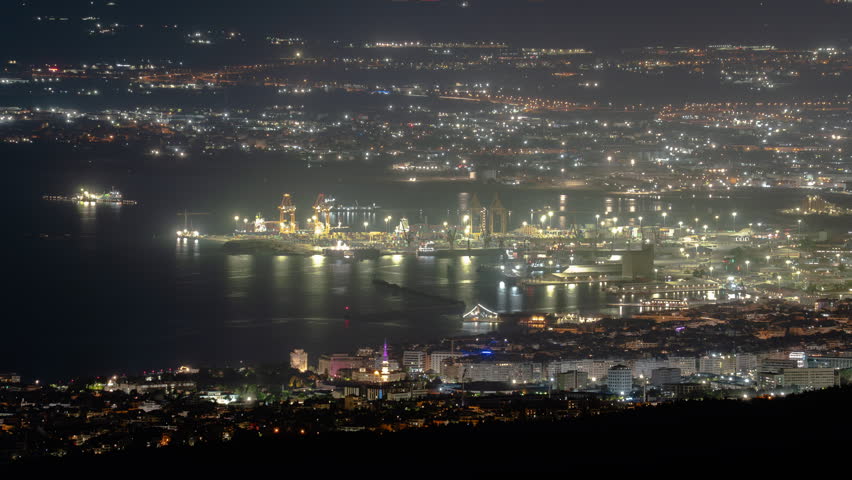 Thessaloniki harbor cranes and waterfront districts glow at night as city lights flicker in a long telephoto timelapse.