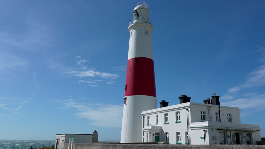 A cinematic shot of the iconic red and white Portland Bill Lighthouse in Dorset, England. This historic maritime landmark stands against a bright blue sky, representing coastal heritage and travel.