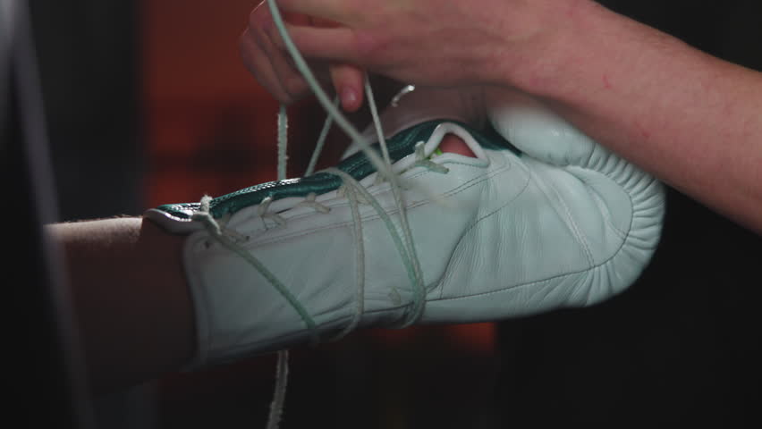 Hands tightening frayed lace on white leather boxing glove with teal trim inside dim gym, showing focused preparation, tactile texture, and worn training gear detail