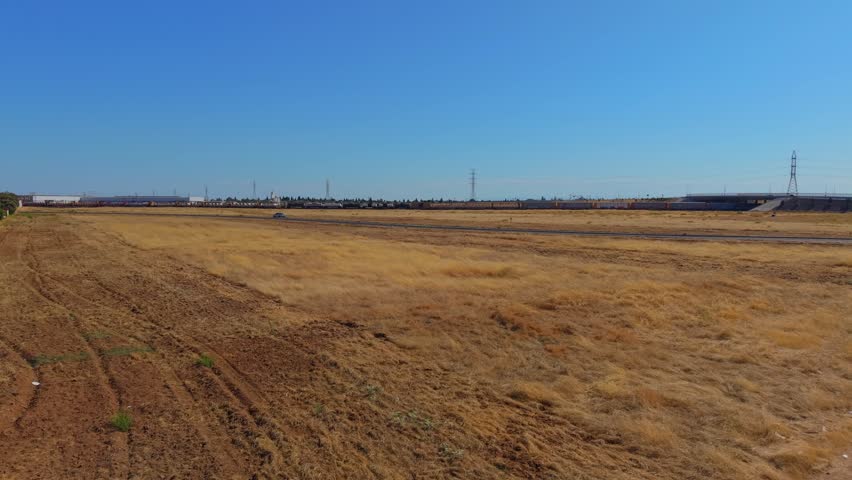 Ascending aerial drone footage rising over a dirt field in Fresno, California, revealing a train actively moving along the railroad tracks below. High voltage power lines and transmission towers stretch across the landscape, with road traffic visible nearby. This dynamic, cinematic aerial reveal captures the region