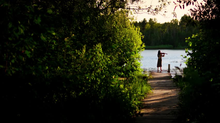 Solitary musician playing violin on tranquil lakeside wooden pier surrounded by lush green forest foliage at golden hour sunset in peaceful nature