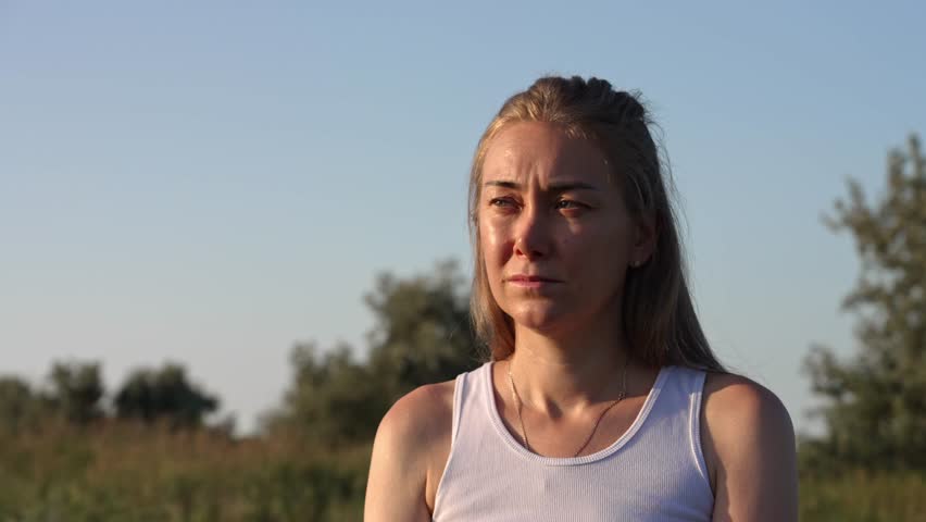 Depressed young woman with tears in her eyes is crying in a field at sunset, her face illuminated by the warm light showing her sadness and despair as she looks away into the distance