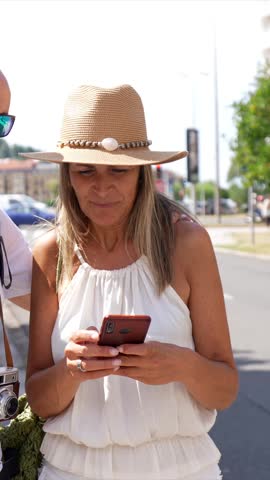 Happy mature couple on vacation using a smartphone map for navigation while exploring a sunny city street. The man, a photographer with a vintage camera, and the woman look for directions together