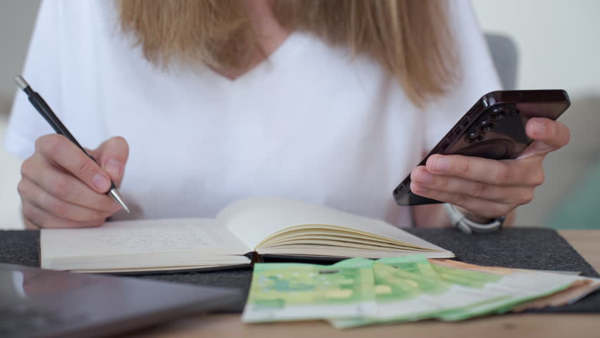 Woman managing home budget, writing notes with phone and euro cash nearby. Concept of household expenses, bills and personal finance.