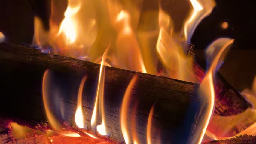 Orange flames flicker around charred wood logs in a cozy indoor hearth with warm lighting.