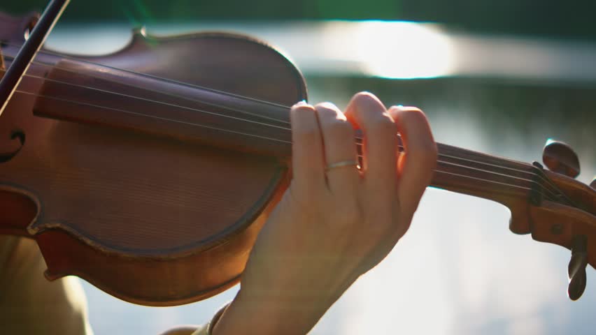 Close-up of a musician hand gracefully playing a wooden violin outdoors near water, capturing delicate strings, bow movement, and warm natural sunlight reflections