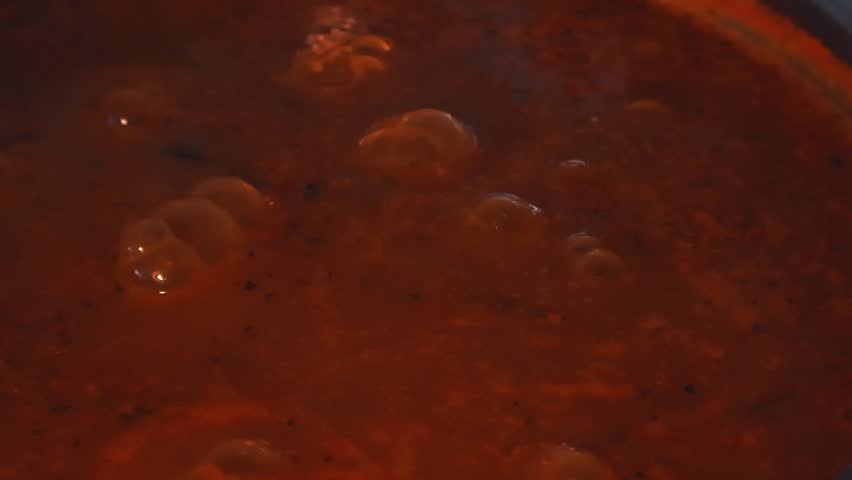 Close up view of thick, red tomato soup boiling slowly in a cooking pot, with large bubbles rising to the surface and bursting, showcasing the texture of the hot, simmering meal