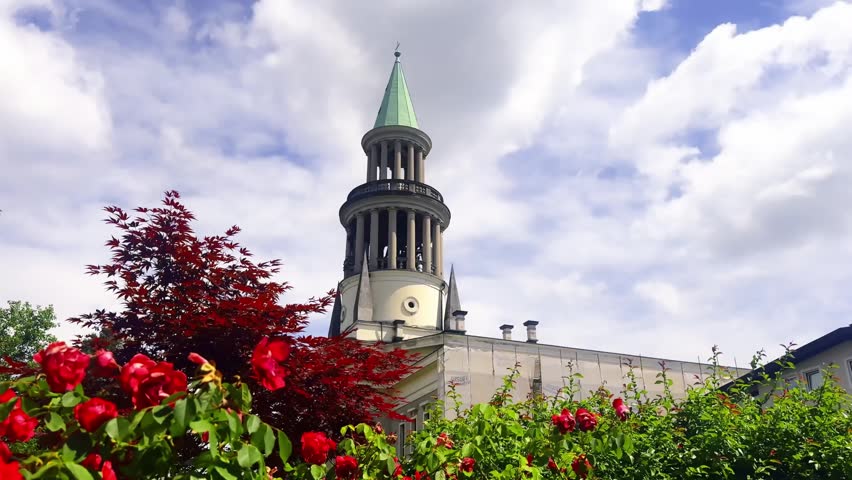 Church tower in Tacen, Slovenia, rising behind red roses and leafy garden plants under bright summer clouds.