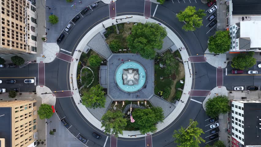 Circular stone monument fountain anchors downtown roundabout in Easton, Pennsylvania, symmetrical green trees frame paved plaza beneath surrounding tall brick buildings. Aerial drone top down rise