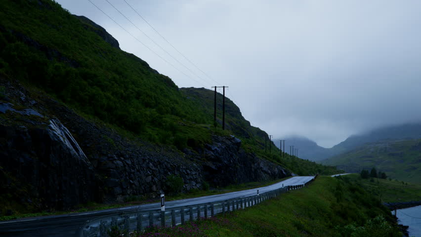 Wet two-lane road curves along a rocky hillside with power lines and low clouds hanging over a Nordic valley.