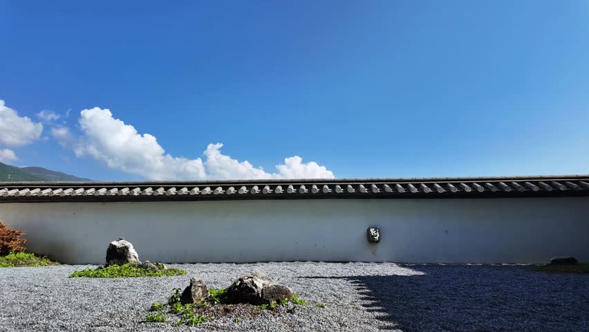 Serene time-lapse video of white clouds drifting rapidly across a clear blue sky above a traditional Japanese garden wall featuring dark grey tile roofing. White plastered wall with gravel ground, scattered rocks and small green plants below. Distant mountains in the background. Captures peaceful passage of time over timeless east Asian architecture on a bright sunny day. Ideal for backgrounds, travel, culture, meditation and nature themed projects.
