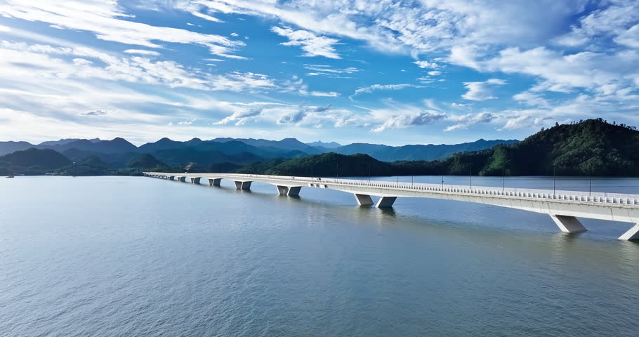 Aerial view of a long concrete bridge spanning a wide lake toward lush green mountains under a bright cloudy sky. Excellent infrastructure background for travel.