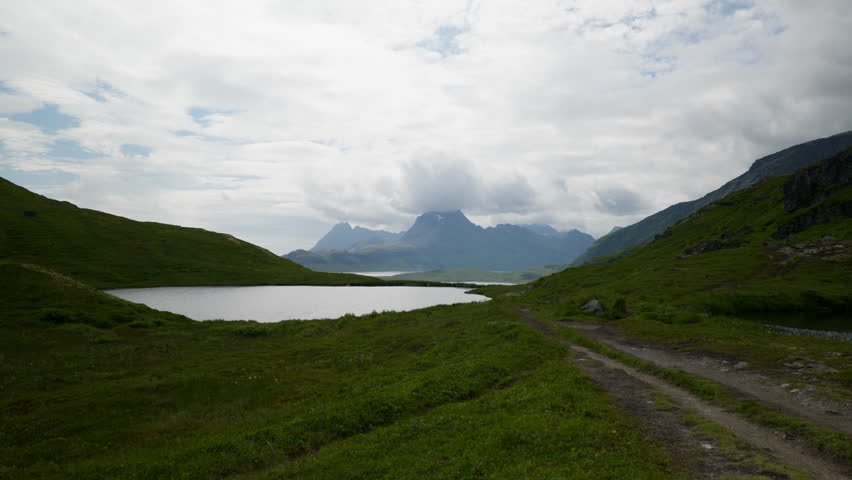 Gravel track follows a small hilltop lake through green Norway Lofoten mountains beneath a cloudy summer sky.