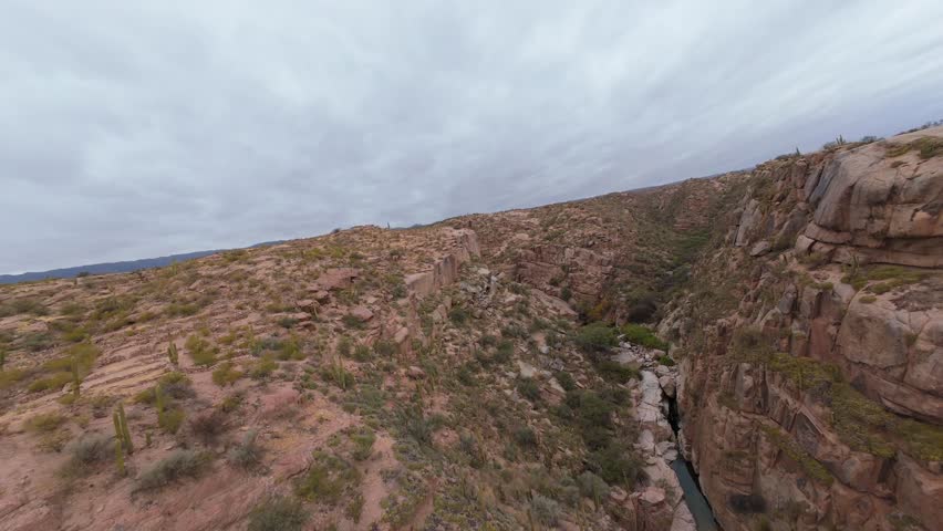 Steep rocky gorge over flowing river stream on a moody, cloudy afternoon. Forward aerial traverse on natural landscape.