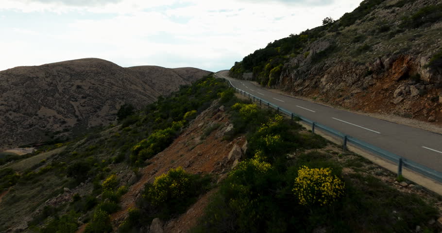 A fast-moving aerial shot follows a winding mountain road through rocky hills, green shrubs, and yellow flowers, with guardrails and sharp curves ahead.
