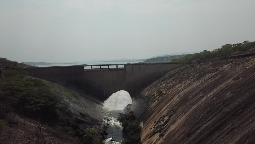 Aerial shot of a large concrete dam with water forcefully discharging from the central spillway, creating a massive white waterfall that crashes into the rocky gorge below, surrounded by natural lands