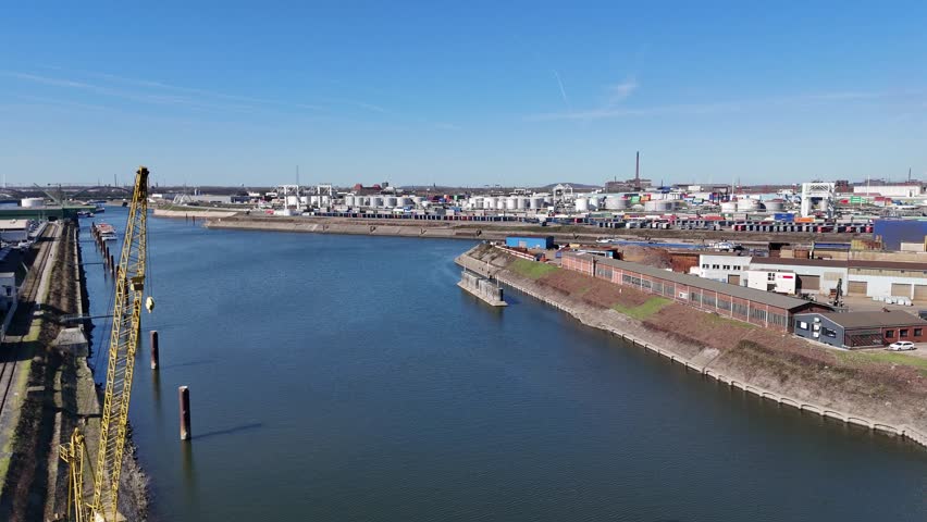 Drone glides past a foreground crane while opening the view into a large harbor basin with tanks and containers.