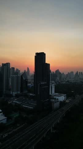 Aerial view of a modern cityscape in Bangkok, Thailand with skyscrapers silhouetted against a vibrant orange and yellow sky as the sun sets behind the buildings, creating a serene urban scenery.
