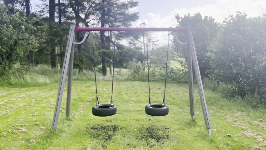 Two tire swings gently moving in the wind, hanging from a swing frame surrounded by green plants, trees and bushes. A peaceful outdoor scene evoking childhood memories and a calm garden atmosphere.