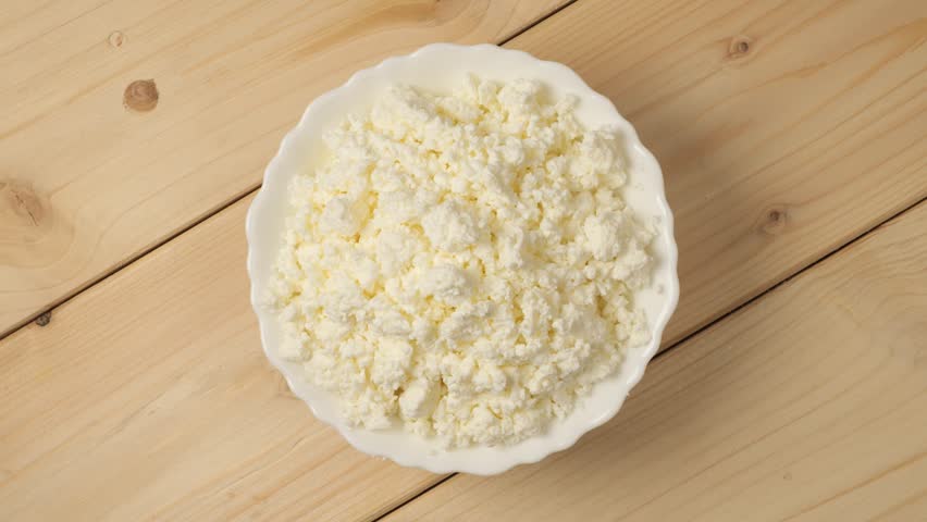 White porcelain bowl with fresh cottage cheese rotating on a wooden table, a healthy breakfast food