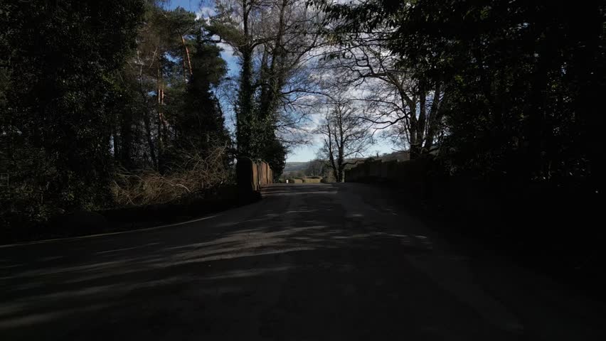 A frontal view of a quiet shaded road leading into Rowsley village, Derbyshire, lined with tall trees and soft afternoon light through branches