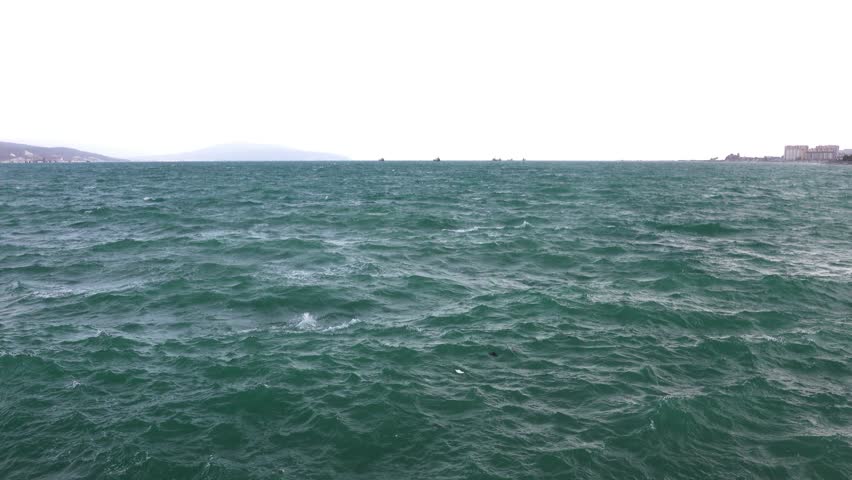 Stormy sea with powerful waves, distant city skyline and ships on the horizon under dramatic cloudy sky.