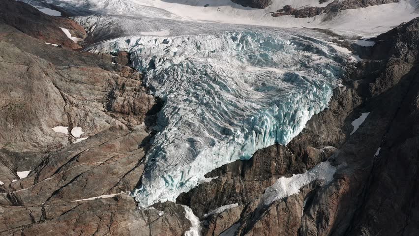 An Aerial view of a large, flowing Trift Glacier in Switzerland, nestled in a deep valley between steep, rocky, gray mountains with patches of snow