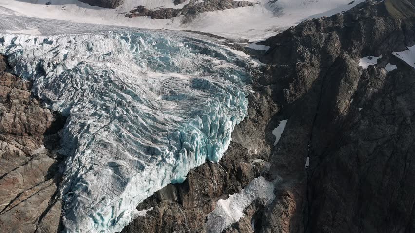 An aerial view of large, flowing Trift Glacier in Switzerland, nestled in a deep valley between steep, rocky, gray mountains with patches of snow
