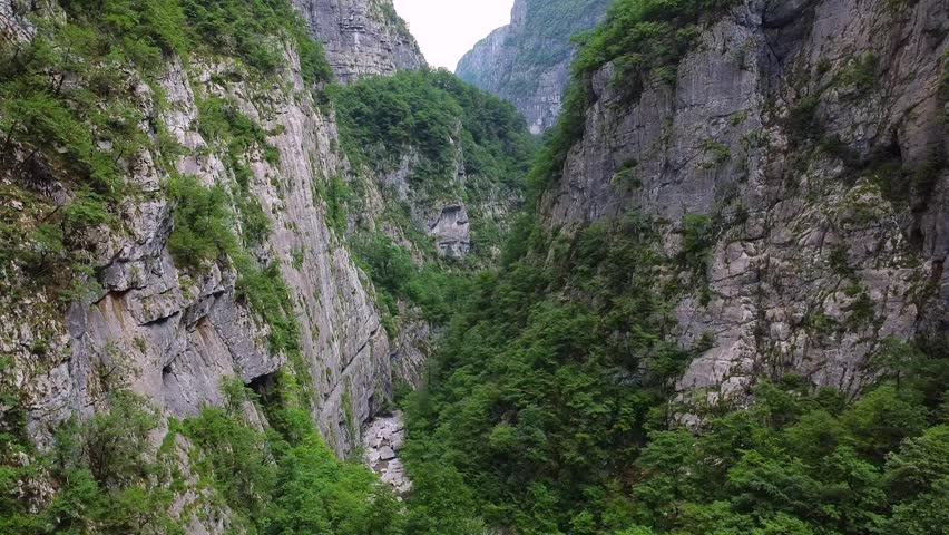 Majestic mountain gorge with steep rocky cliffs covered in lush green trees. A narrow, winding riverbed carves a path through the bottom
