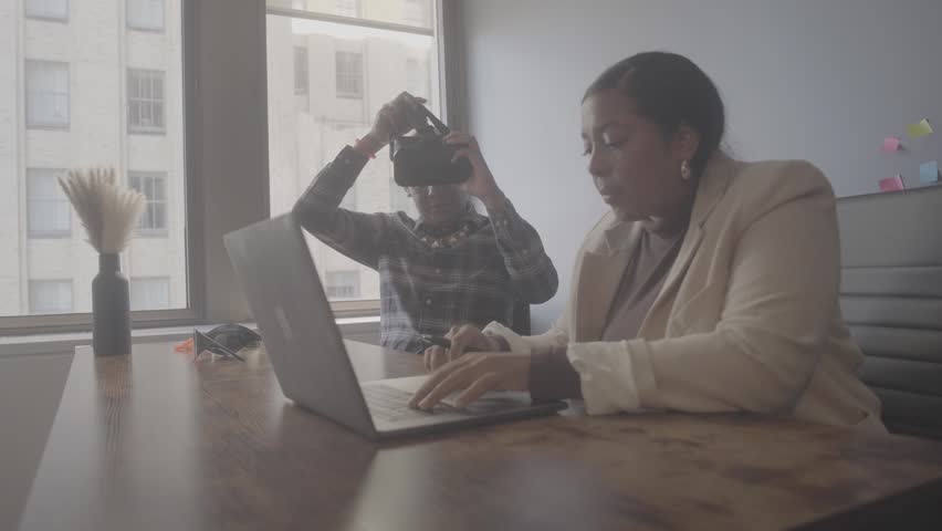 Two people are working in an office. One person is using a laptop while the other is trying on virtual reality glasses. They focus on their tasks in a bright space with large windows.