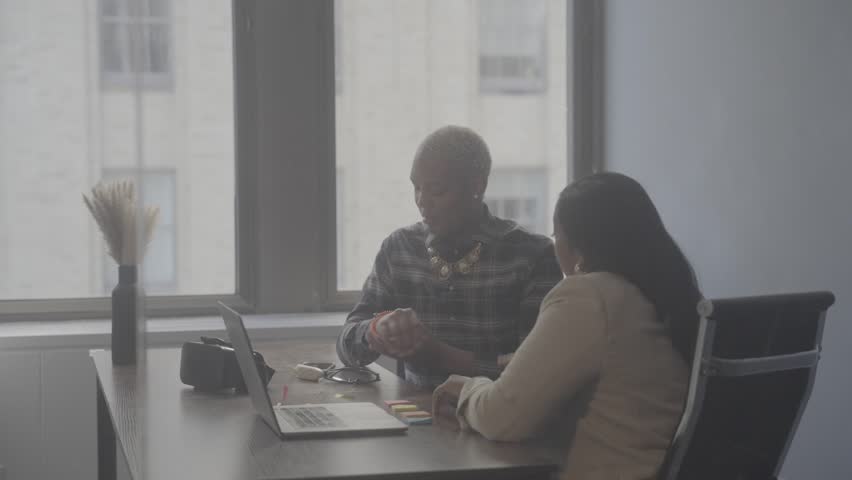 Two people are sitting at an office table discussing their plans while using a laptop. Sunlight comes through the windows as they engage in conversation.