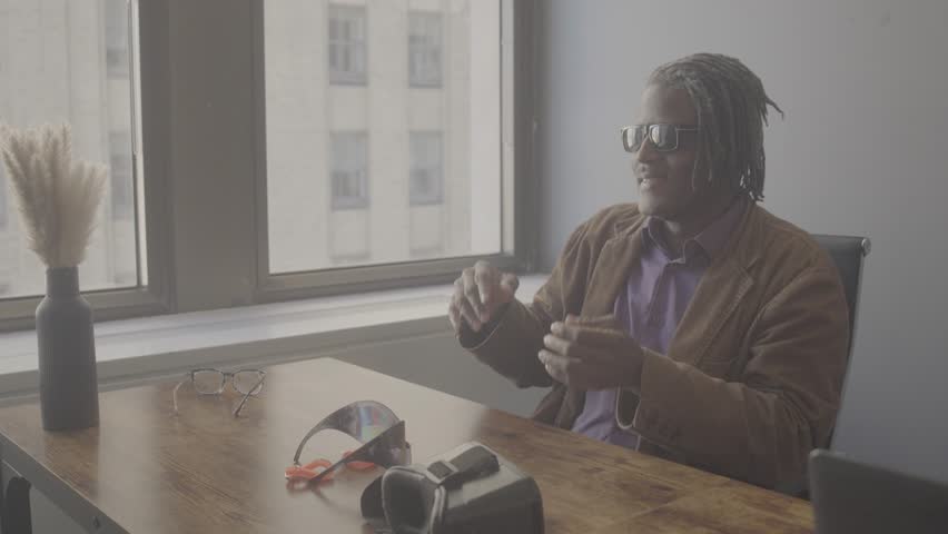 A man sits at a desk in an office by a large window. He waves his hand while speaking. Objects like glasses and scissors are on the desk. The room gets natural light.