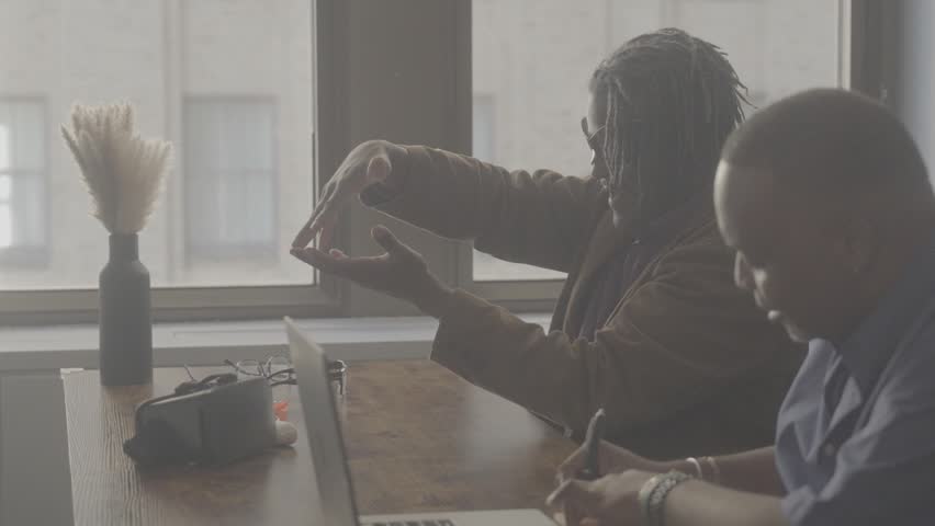 Two people communicate at a table in a well-lit room. One person gestures while the other listens and writes. They appear to be focused on a discussion.