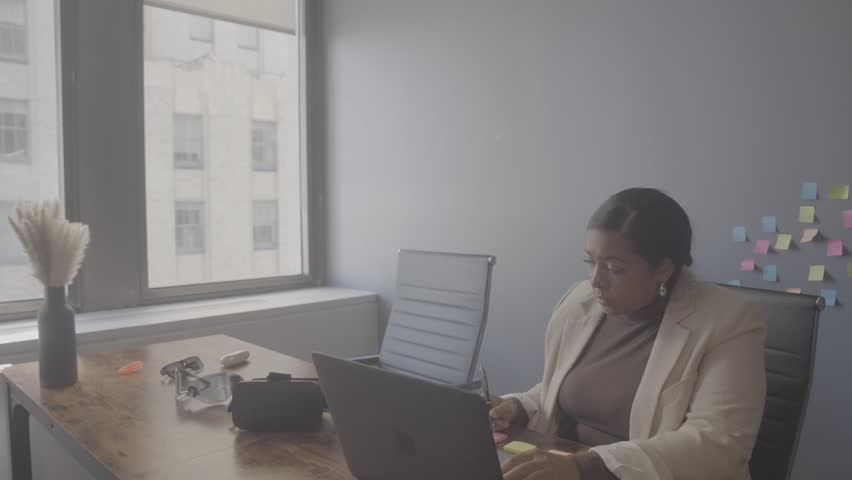 A person sits at a desk with a laptop and notepad in an office. Sticky notes are on the wall. Bright sunlight comes through the window.