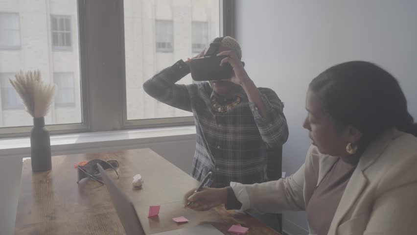 Two people are in an office during the day, with one using virtual reality gear while the other watches. They are engaged in a technology session with notes on the table.