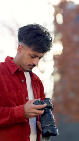 Young Moroccan male photographer looking at his professional digital camera and taking pictures outdoors. Focused creative artist at work. Vertical