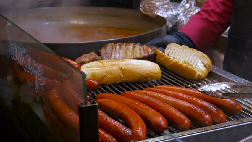Street vendor grilling sausages and hot dog buns at market stall. Close-up of a street food vendor grilling delicious sausages, hot dog buns, and meat at an outdoor festival or market